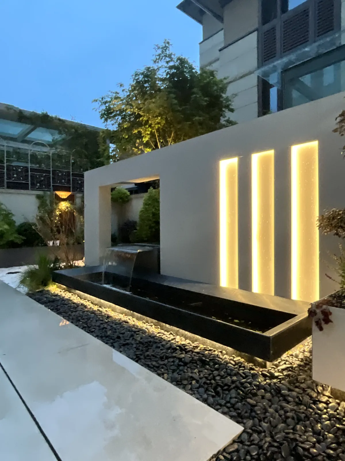 郑州兰乔圣菲完工实景:从另一端回看流水景墙,水面与铺装映出傍晚灯光。